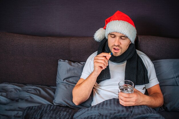 Sick young man in Christmas red hat sits on the bed