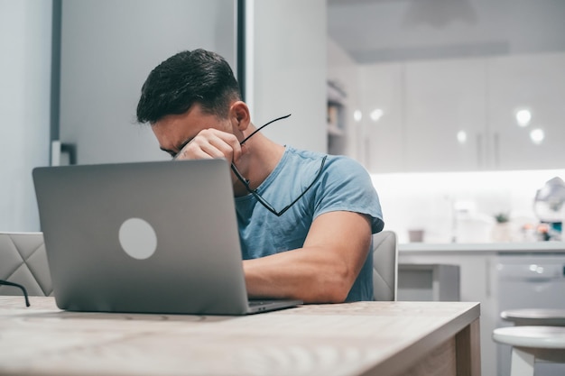 Sick young man feeling unhealthy at home working with laptop on the table alone Teenager boy taking off his glasses after tired day Overwork concept