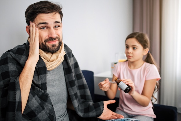 Sick young man look on camera. He hold hand on head. Guy covered in blanket. Small girl pour some syrup into spoon.