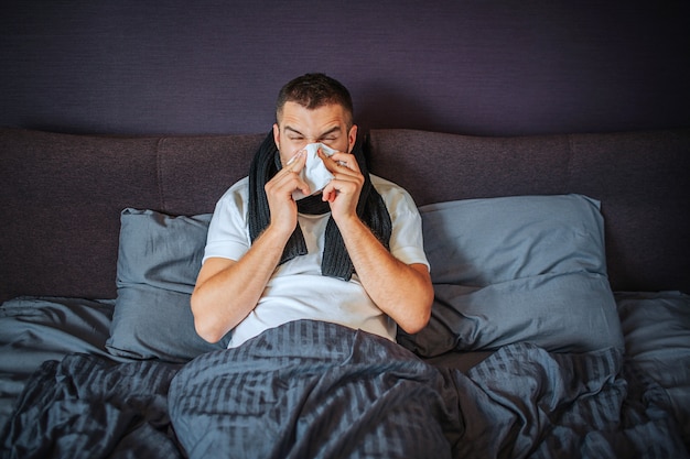 Sick young man sits on bed. He is covered with blanket. Guy sneezing into tissue. He suffers. Young man feels terrible. He is concentrated on sneezing.