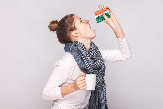 Sick young woman holding cup with tea, many pills and antibiotics. Studio shot, isolated on gray background