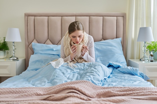 Sick young woman at home in bed with cup of hot drink and handkerchief