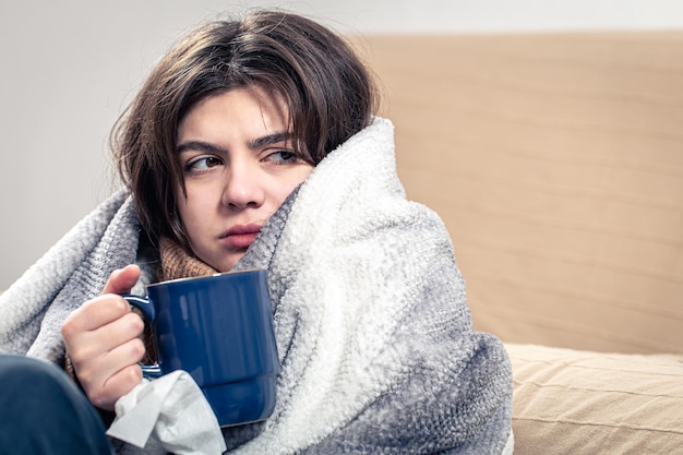 A sick young woman sits with a cup of tea wrapped in a blanket