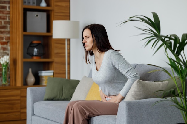 Sick young woman suffering from menstrual pain Woman with hands squeezing belly having painful stomach ache or period cramps sitting on sofa Abdominal pain gastritis and painful periods concept