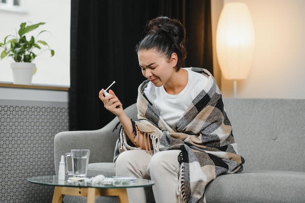 Sick young woman in warm clothes spraying throat while sitting on couch at home