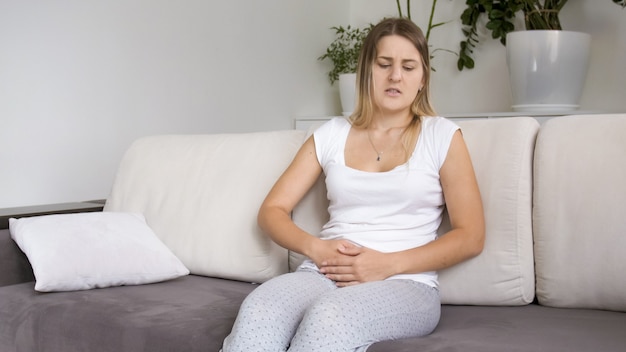 Sick young woman with abdominal or stomach pain sitting on sofa in living room