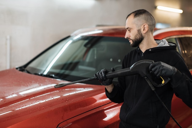 Side angle view of a man worker cleaning modern red automobile with high pressure water at car wash