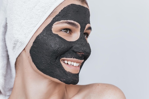 Side closeup view of beautiful smiling young female doing beauty treatment on her face clean skin with black organic clay looks aside with toothy healthy smile Woman applying natural facial mask