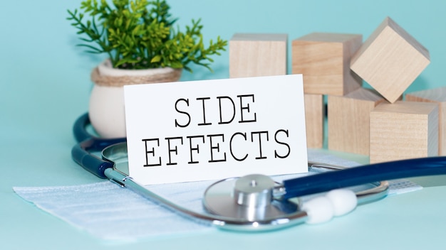 Side effects - words written on white medical card, with\
medicine mask, stethoscope, green flower and wooden blocks on\
background.