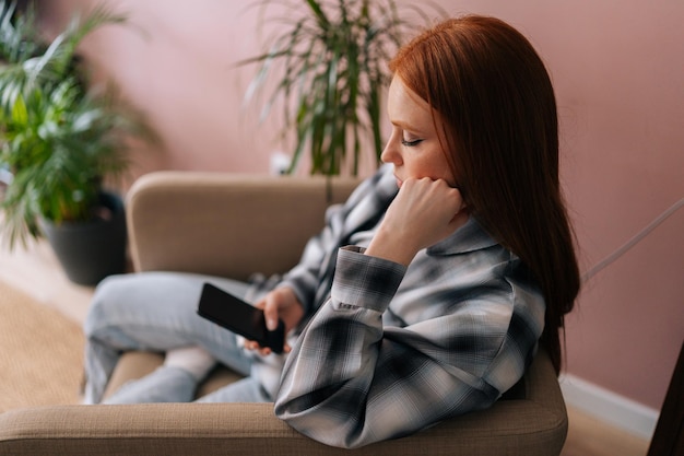 Вид сбоку на скучающую красивую рыжую женщину-пользовательницу смартфона женщина пишет текстовое сообщение в чате, печатая сидя в кресле с помощью приложения для покупок