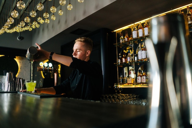 Side lowangle view of barman pouring fresh alcoholic drink into\
glasses with ice cubes behind bar counter in modern nightclub