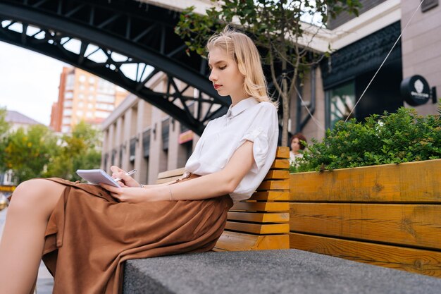 Side lowangle view of thoughtful young woman resting on bench in city street writing plans for trip in notebook Portrait of pretty lady creating drawings