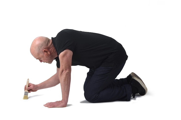 side of a man on knees painting on the floor with a brush on white background