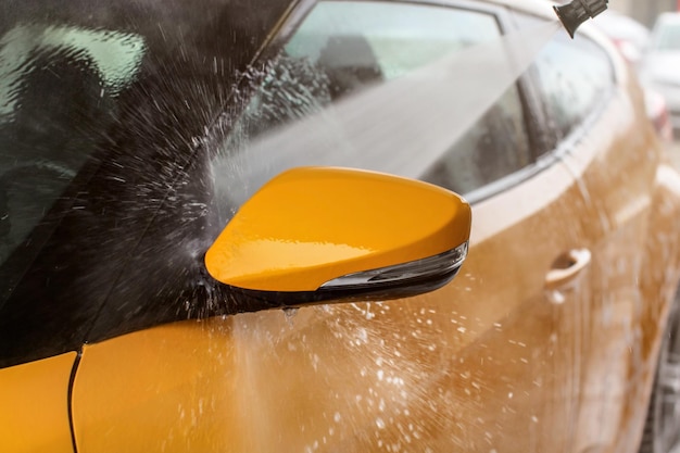 Side mirror of yellow car washed in self service carwash, jet spray of water hitting window water drops flying to all sides.