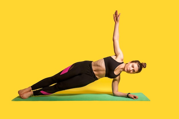 Поза боковой планки. Молодая фитнес-женщина в обтягивающей спортивной одежде практикует йогу, делает упражнения васиштхасаны с поднятой одной рукой, тренирует мышцы. студийный снимок, спортивные тренировки на желтом фоне