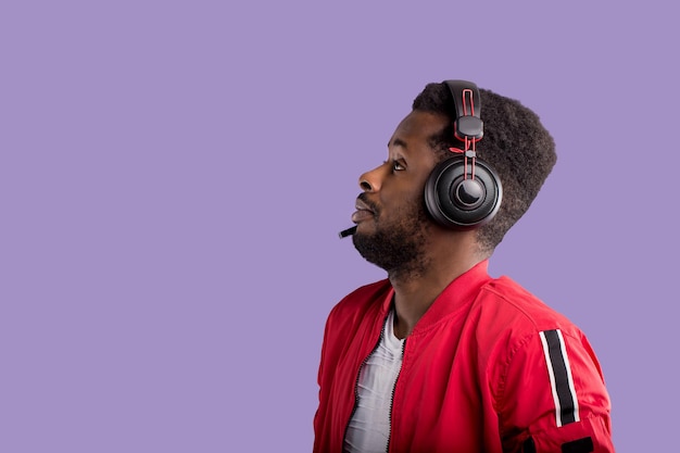 Side portrait of a beautiful afroamerican men in a red sports jacket and headphones on purple background with copy space. Music, DJ concept