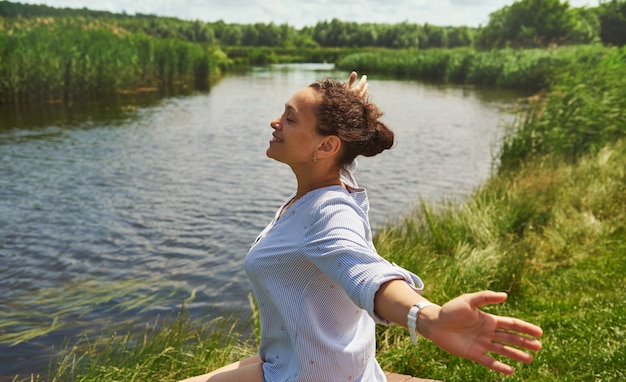 Боковой портрет счастливой и улыбающейся женщины с протянутыми руками, наслаждающейся красотой природы на фоне реки