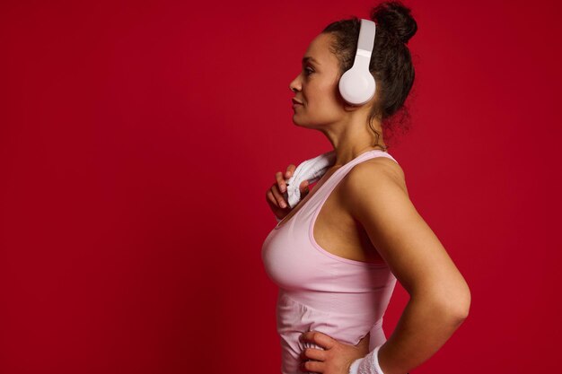 Side portrait of a Latin American sporty woman with headphones and terry towel on her shoulders isolated over red colored background with copy ad space