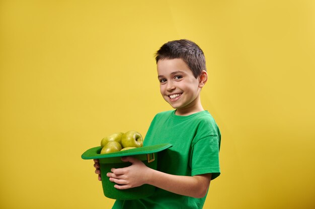 Side portrait of a smiling boy holding a leprechaun hat full of\
green apples