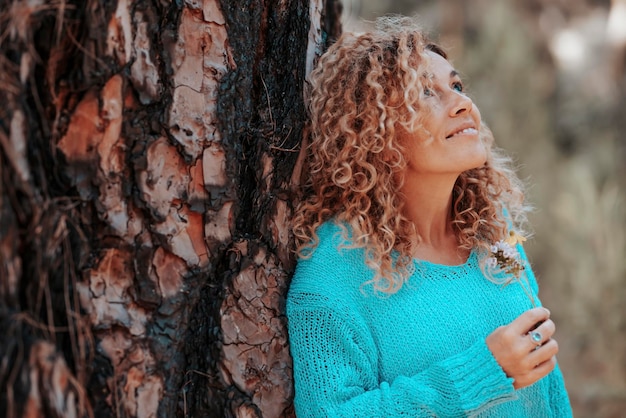 Side portrait of woman smiling and enjoying nature in outdoor leisure activity Female people tourist against a tree trunk smile and admire nature environment around Concept of travel and happiness