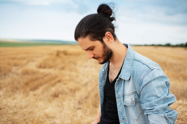 Side portrait of young guy in denim jacket with ponytail hairstyle in hay field.