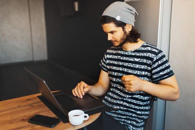 Side portrait of young handsome man working home on laptop; coffee cup and smartphone on wooden table.