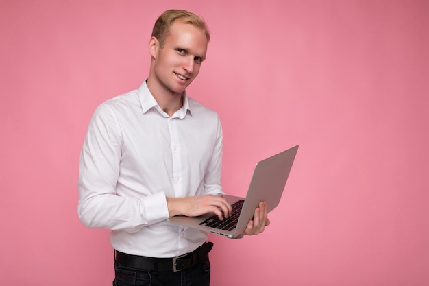 Side profiel foto shot van knappe glimlachende zelfverzekerde blonde mannelijke persoon met computer laptop typen op toetsenbord dragen wit overhemd kijken camera geïsoleerd over roze achtergrond