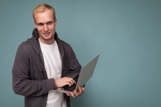 Side profiel foto shot van knappe zelfverzekerde blonde man met computer laptop typen op toetsenbord met wit t-shirt en grijze trui kijken camera geïsoleerd over blauwe achtergrond. Lege ruimte