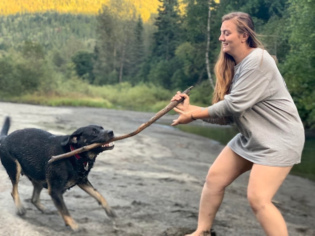 Photo side profile of a woman and dog playing tug of war with a large stick on a riverbed