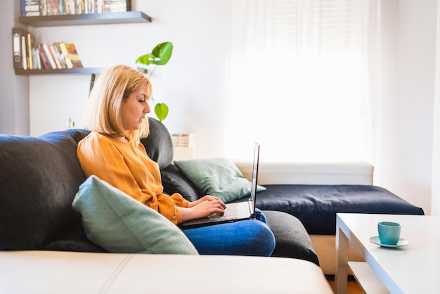 Photo side shot of adult woman in jeans and blouse sitting on sofa in living room browsing netbook