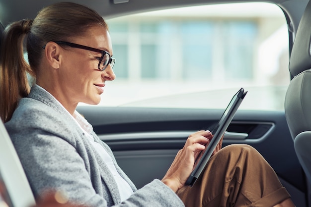 Side of a successful smiling business woman wearing eyeglasses using digital tablet, working while sitting on back seat in the car, business trip. Transportation and vehicle concept