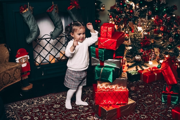 Side view of adorable little girl with tails in white sweater looking at beautifully decorated Christmas tree with christmas balls