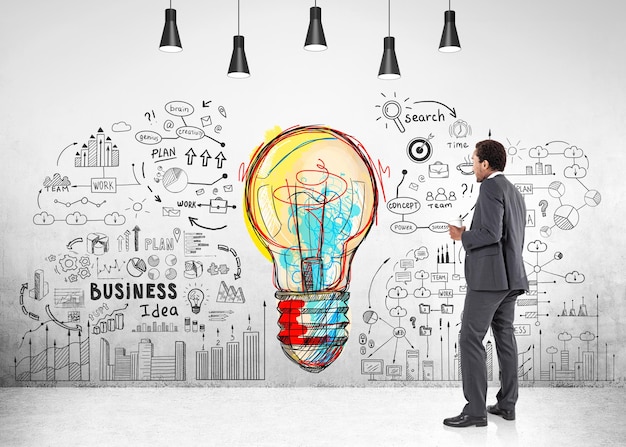 Photo side view of an african american businessman in a dark gray suit standing and holding a coffee to go near a concrete wall with a colorful light bulb sketch and business icons.