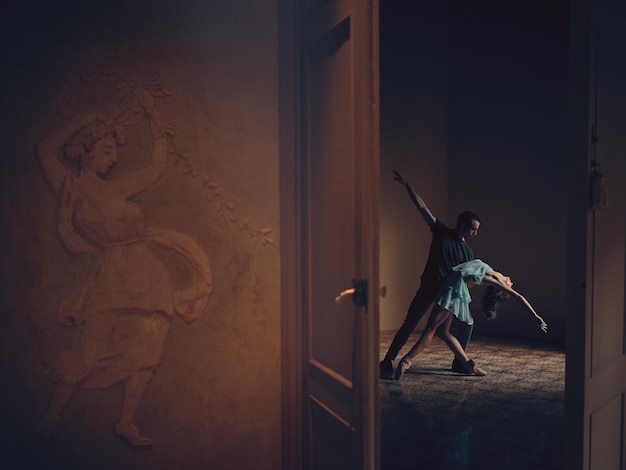 Side view of anonymous man and tender ballerina dancing in empty room behind opened door in dim light