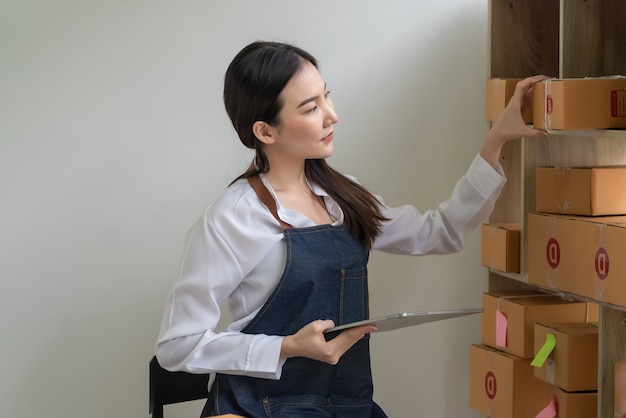 집에서 고객에게 배달하기 전에 소포 상자를 확인하기 위해 태블릿을 들고 있는 아시아 여성 온라인 비즈니스 소유자의 측면 보기.