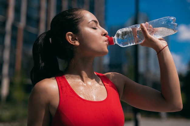 Side view of athletic sweaty young woman with brown wavy ponytail wearing red sports bra and drinking water from plastic bottle