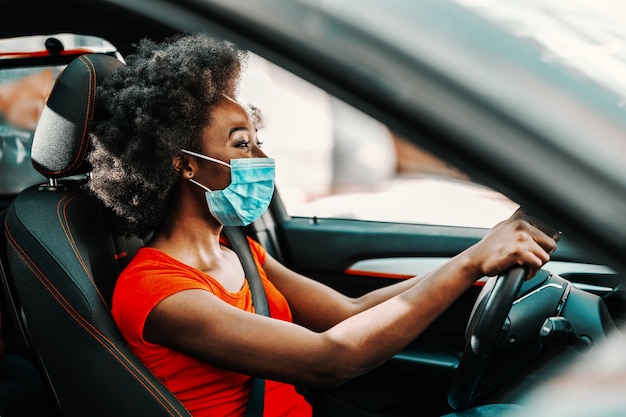 Side view of attractive african woman with short curly hair with face mask on sitting and driving car. Prevention from spreading corona virus / covid 19 concept.