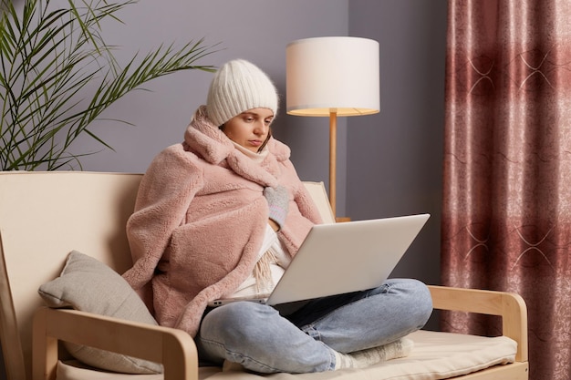 Side view of attractive frozen woman being in cold room sitting\
on the cough wearing warm hat mittens and cap getting warm in fur\
coat while working online at home