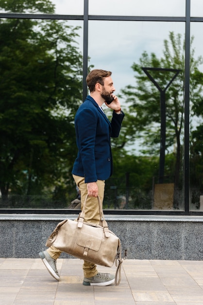 Side view of an attractive smiling young bearded man wearing jacket walking outdoors at the street, carrying bag, talking on mobile phone