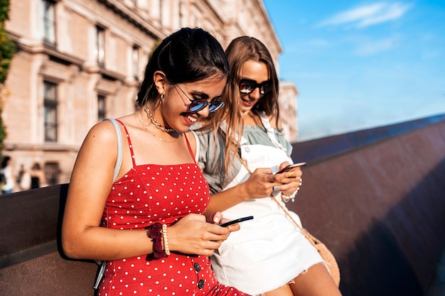 Side view of attractive young female friends using mobile phones while hanging out on city street during summer