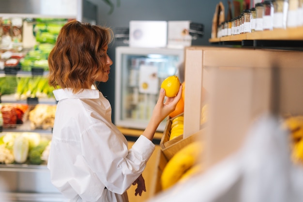 Side view of attractive young woman choosing fruits for diet checking lemon in grocery store at the