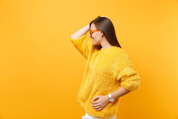 Side view of attractive young woman in fur sweater, orange glasses putting hand on head isolated on bright yellow background. people sincere emotions, lifestyle concept. copy space for advertisement