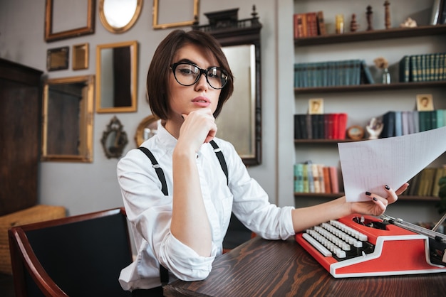 Side view of authoress in glasses and white shirt sitting by the table with paper and typewriter