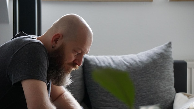 Photo side view of bearded bald man sitting on sofa at home