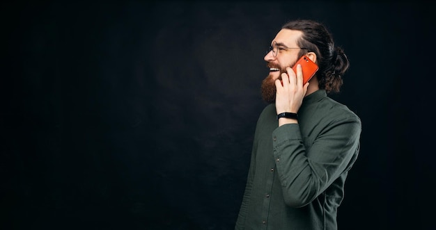 Вид сбоку на бородатого мужчину в очках, который разговаривает по телефону, держа его у уха Студия снята на черном фоне