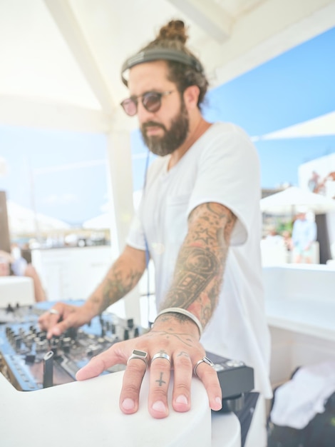 Side view of bearded tattooed disc jockey wearing white t shirt and silver accessories playing music while standing at party zone of hotel