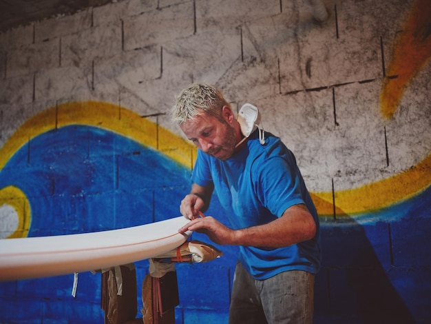 ワークショップで塗装する前にサーフボードのに粘着剤の薄いテープを接着するひげのタトゥーされた男のサイドビュー