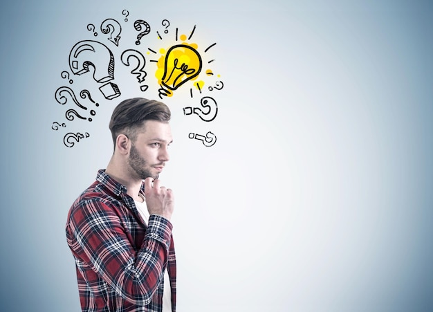 Photo side view of a bearded young man in a checkered shirt and a white t shirt thinking near a gray wall with many question marks and a yellow light bulb drawn on it. mock up