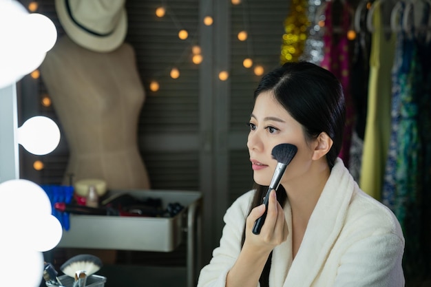 side view of beautiful chinese host doing make up in backstage looking in mirror. young asian entertainer woman applying dry cosmetic tonal foundation on the face using makeup brush in dressing room.