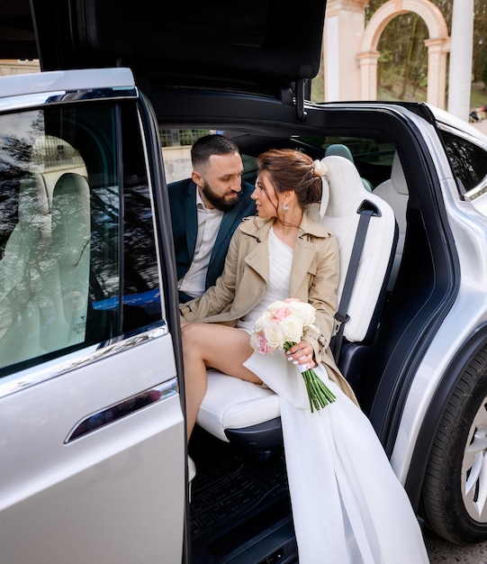 Side view of beautiful couple wearing in trendy outfits sitting in luxury car with opened upper doors smiling and looking to each other